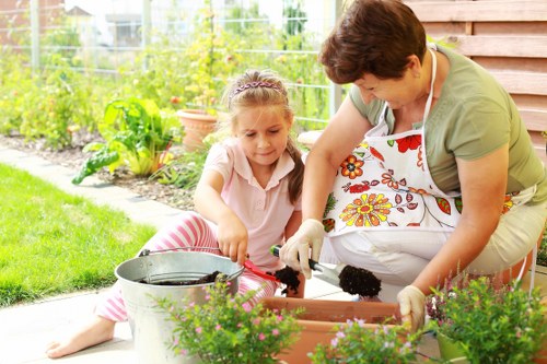 In a well-maintained garden, a woman and a young girl are engaged in planting activities. The woman, wearing a floral apron, is kneeling on the patio, handling dark soil or compost in a rectangular planter box, while the girl, dressed in a light pink top and striped trousers, sits on the edge of the paving slabs, assisting with planting or watering. The foreground features the planter filled with dark soil, accompanied by vibrant pink flowering plants in the near foreground. Behind them, the garden displays lush, green grass, bordered by a tidy flower bed with small shrubs and flowering plants, and a background of leafy trees and a wire fence. The scene is outdoors on a bright, sunny day with natural light illuminating the garden, which appears to be a typical residential back yard in Ealing, with well-kept greenery and a wooden fence providing privacy. Gardening tools and watering cans are nearby, suggesting ongoing garden maintenance, connected to the services provided by Gardening Services Ealing, which specialises in lawn care, planting, and outdoor landscape management.