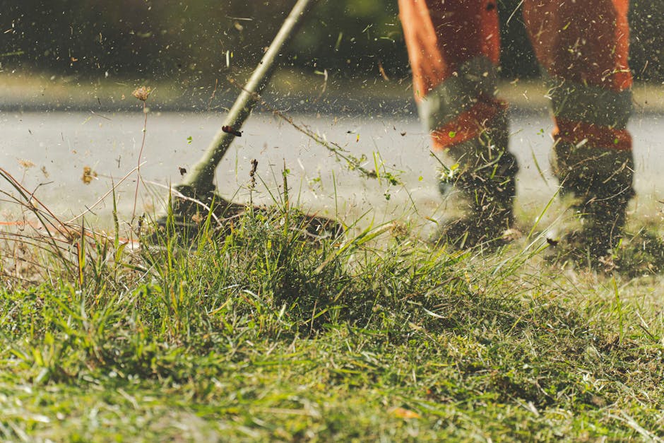 A gardener wearing orange work trousers and sturdy black boots is engaged in garden maintenance, using a rake and a garden fork to clear a patch of overgrown grass, weeds, and fallen leaves from a law